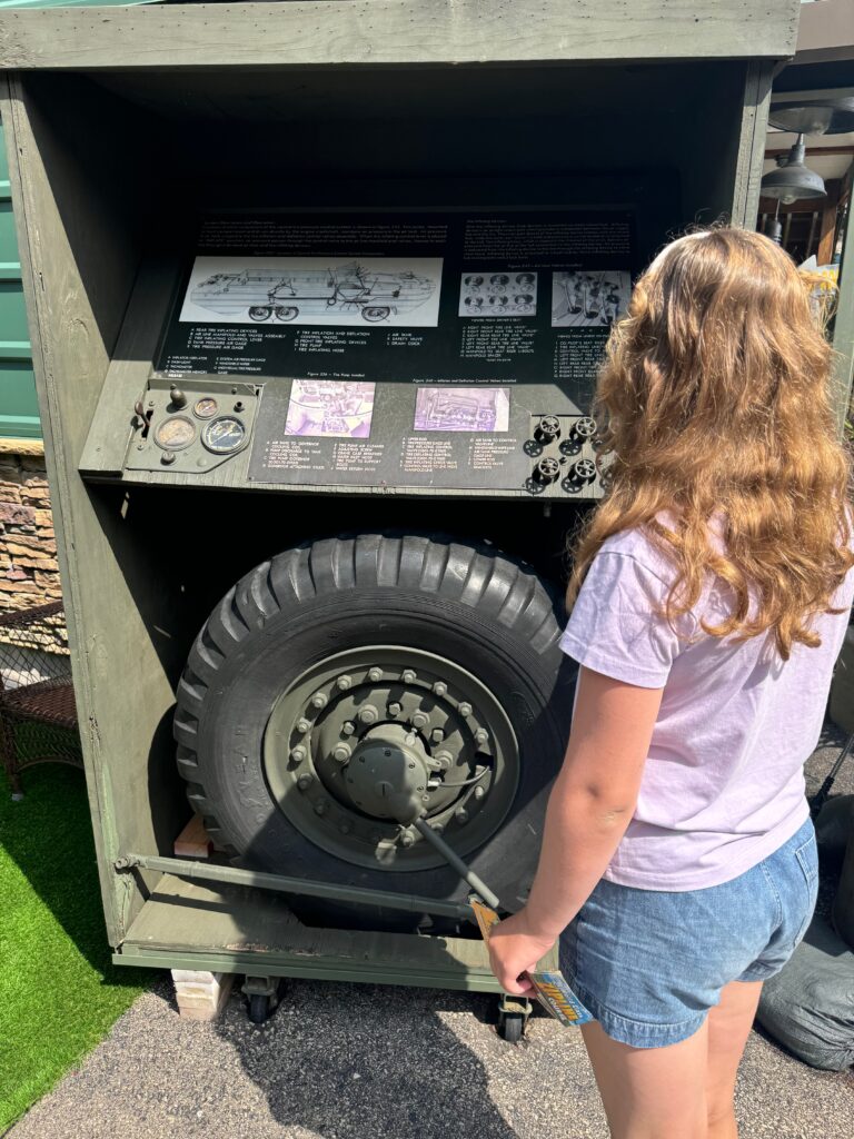 A young girl with blond hair stands facing a display.  The display has a tire from a duck boat as well as other military type controls from the duck boat. 