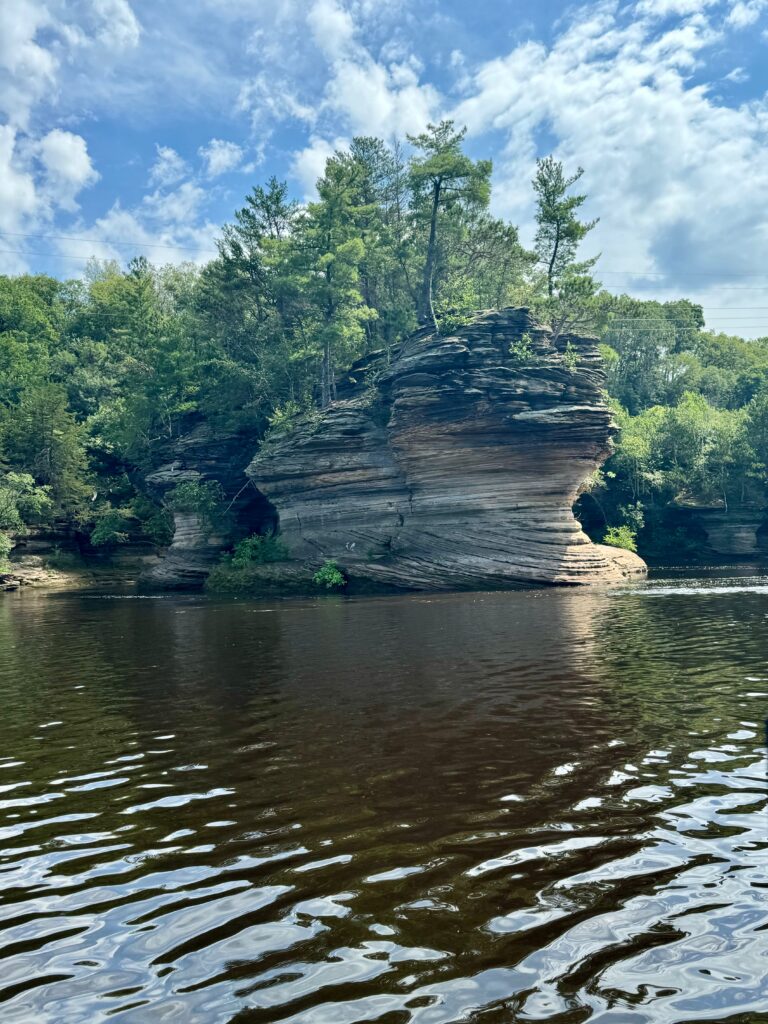 The Wisconsin Dells  rock formation.  One of the large, layered rocks is in the river with a forest behind it.  
