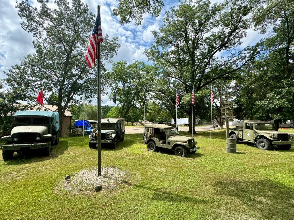 Vintage military vehicles are parked on a grassy area.  A flag pole with the American Flag is in front of them. 