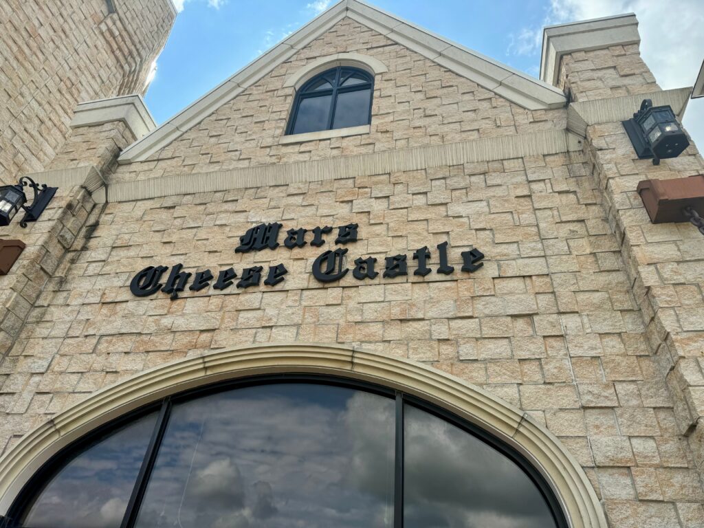 A light brick building stands tall.  There is a small arched window at the top and a larger arched window at the bottom.  In an olden time font reads, "Mars Cheese Castle".  