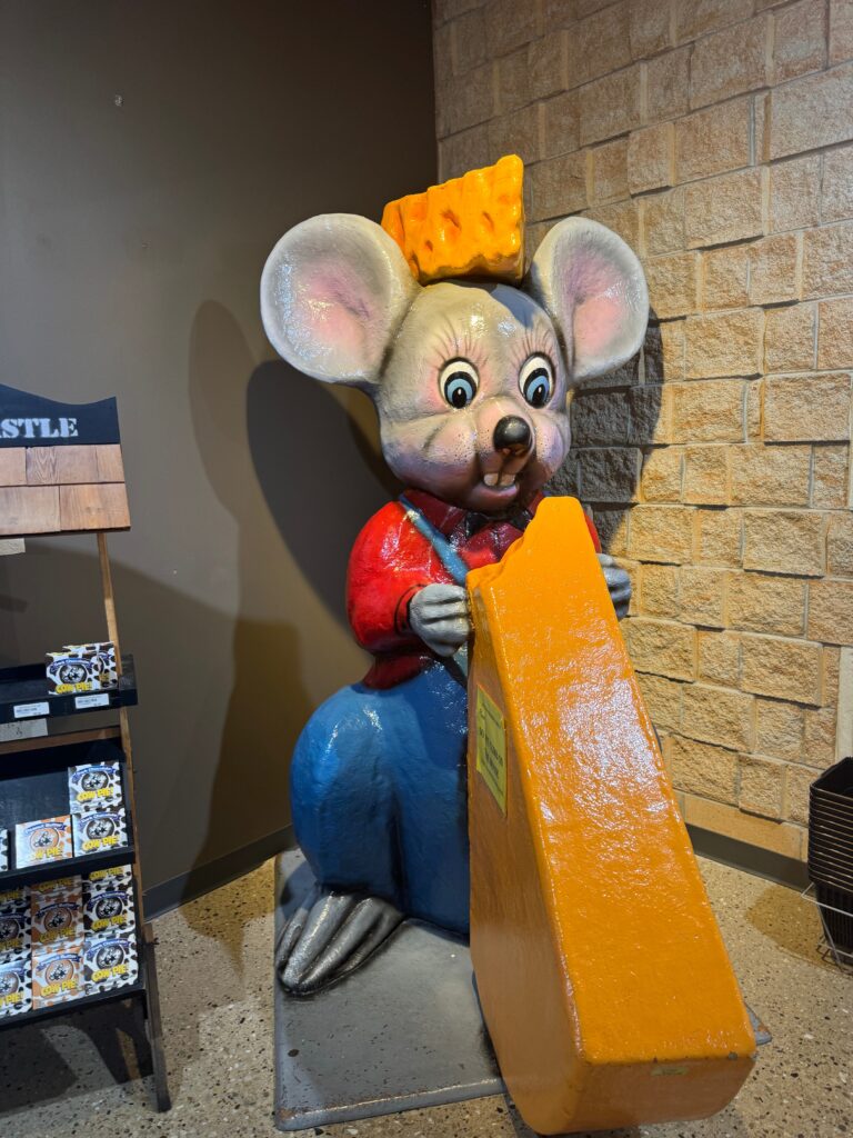 An approximately 7 foot tall mouse statue is inside of a store.  It holds a large triangle of orange cheese and has a cheese hat.  The mouse is wearing a red shirt and overalls.  