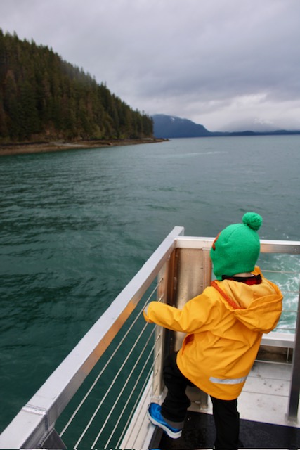 A young child in a yellow rain slicker and a green beanie hangs on to the railing on the stern of a boat.  They have one foot on the bottom railing. Water and a forested mountain lies beyond. 