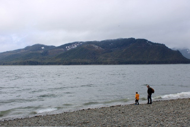 Icy Strait Point Beach- A rocky beach with a child and adult in jackets walking near the shoreline.  A mountain is in the background behind the ocean. 