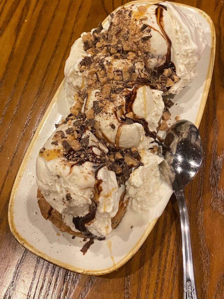 A platter of cake with scoops of vanilla ice cream and drizzles of chocolate and caramel sauce is on a table. A silver spoon rests on the platter.
