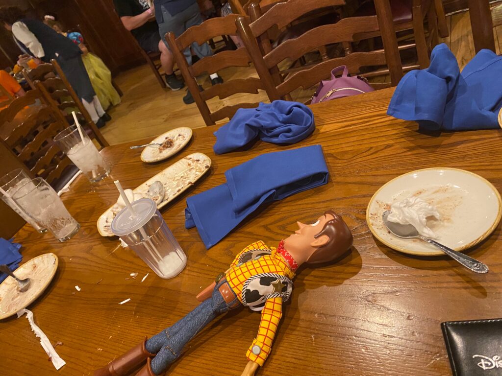 The aftermath of a meal at Liberty Tree Tavern.  Royal blue cloth napkins lay on a wooden table with empty cups and plates.  A Toy Story Woody doll lays forgotten across the table. 