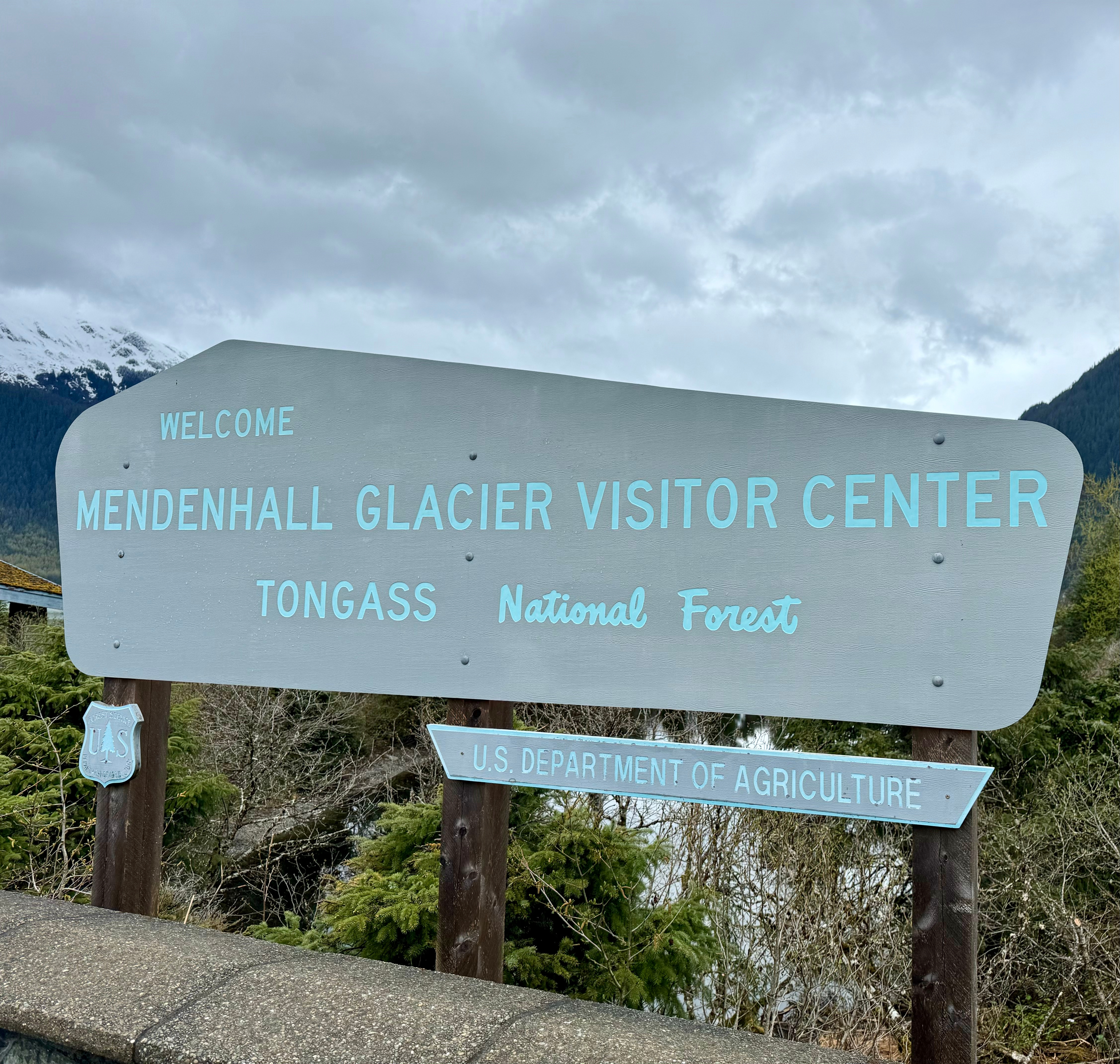 A large grey sign reads, "Welcome Mendenhall Glacier Visitor Center Tongass National Forest" in light blue lettering.  Bushes are behind it. 