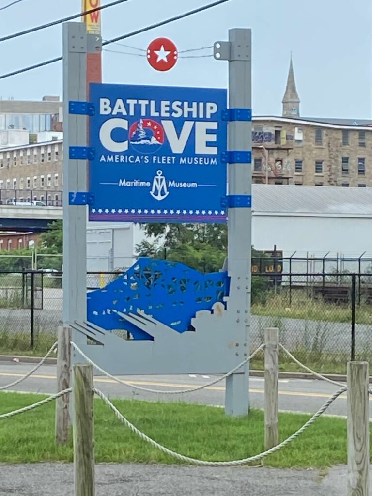 A royal blue sign reads "Battleship Cove"  The 'O' in the word cove is a different design.  The top is red with four white stars and the middle of the 'O' has a battleship going through the water.  The sign is hung with blue brackets on two grey posts.  buildings are in the photo background, including a church steeple.  