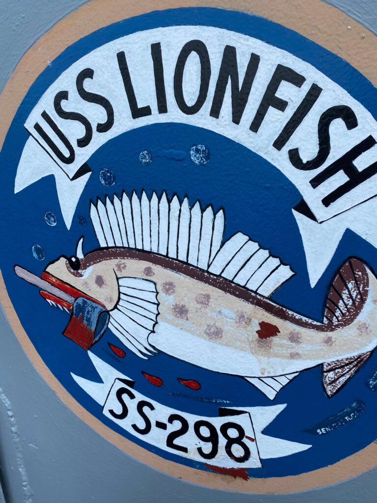 The crest of the USS Lionfish- it is a royal blue circle with a white banner with USS Lionfish in black capital letters.  A lionfish is the center with another white banner, reading SS-298 at the bottom. 