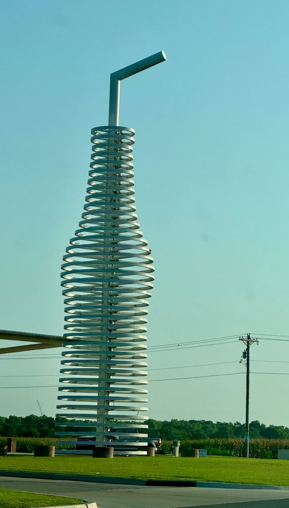 A giant soda bottle, with a straw in it towers over the landscape.  It is not made to look solid, but rather lines and lines making the soda bottle outline. 
