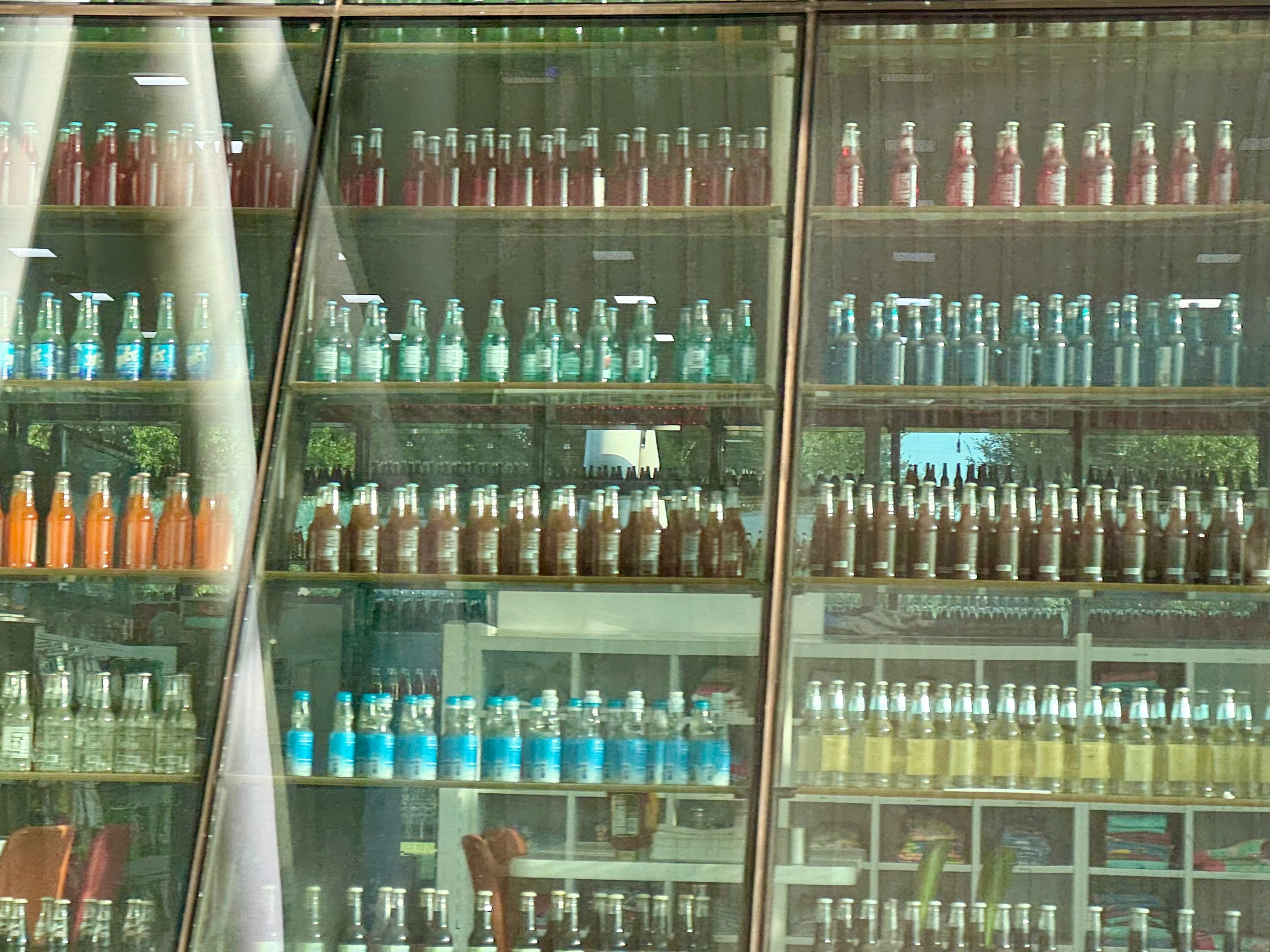 Behind glass are rows upon rows of old fashioned soda bottles arranged by color. 