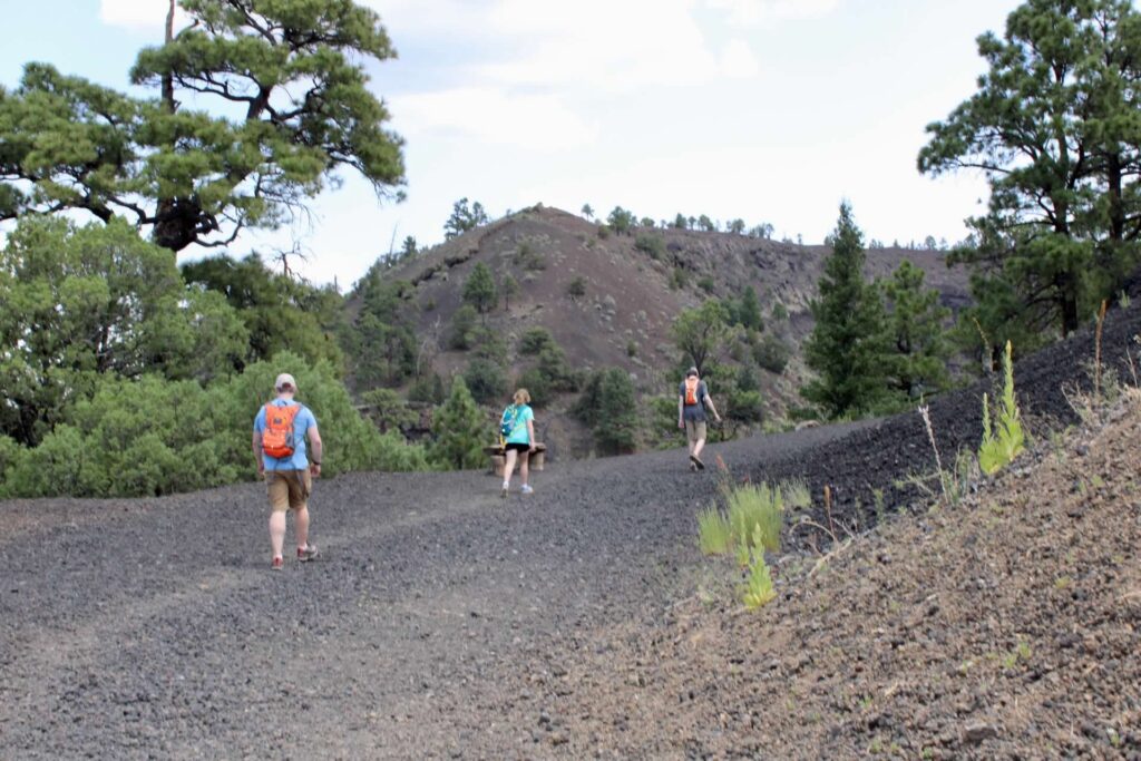 Three people hike along a dirt path. A mountain that looks like it is covered in dirt is in the distance.  Tree dot the area. 