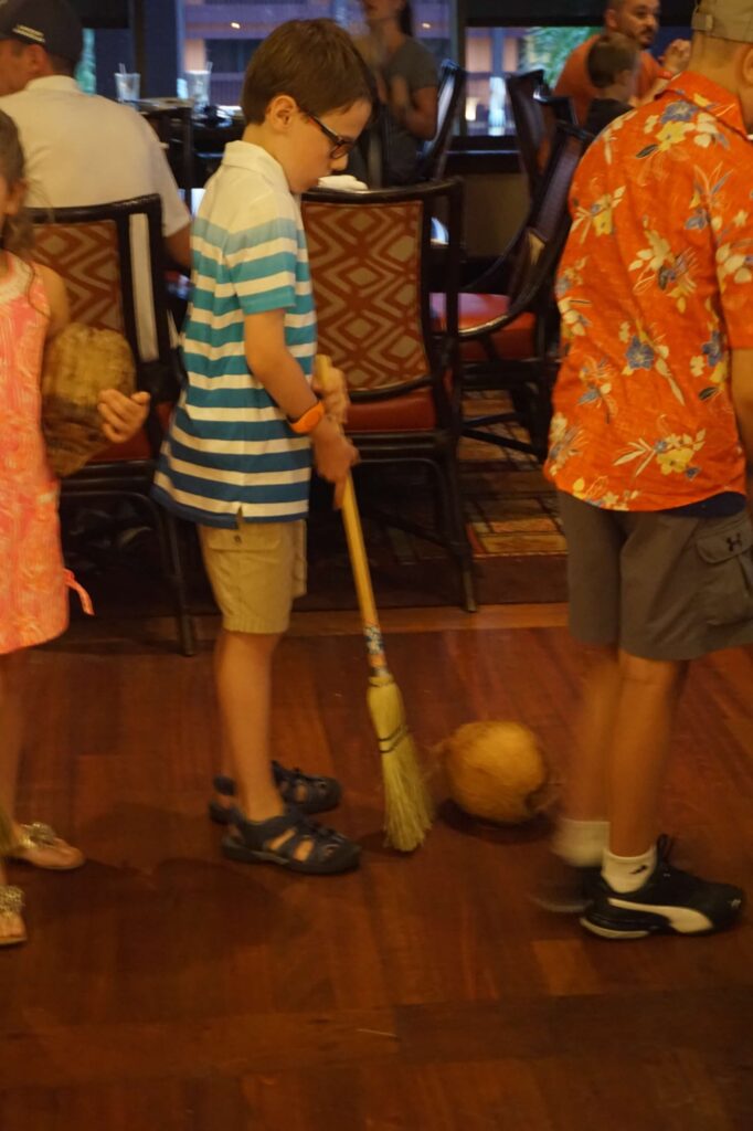 A child pushes a coconut with a broom along the floor of a crowded Ohana restaurant. 
