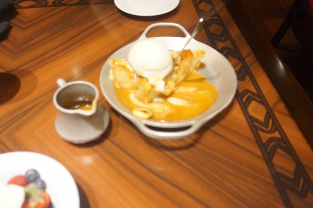 A bowl of Ohana bread pudding sits on a table.  The pudding has a scoop of melting vanilla ice cream on top.  There is a small pitcher of caramel sauce next to the bowl. 