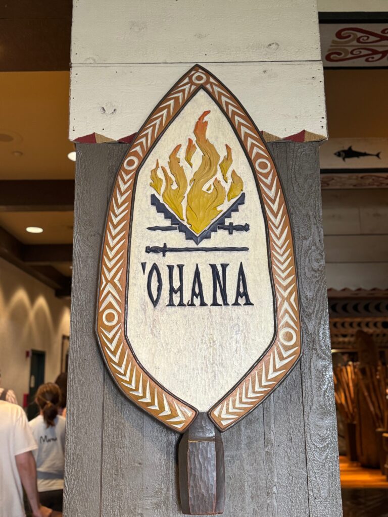 A wooden plaque hangs on a post.  It says "Ohana" and has flames above the word. 