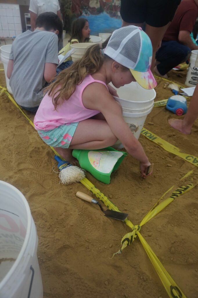 A child is squatted down in sand and appears to be digging.  There is caution tape marking a small square that the child is inside of.  A white five gallon bucket is next to the child. 
