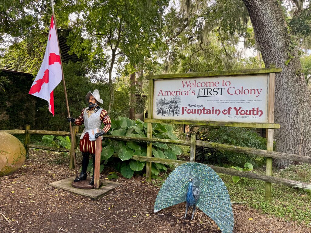 A mannequin in a suit of armor holds a white and red flag.  He is placed next to a sign reading, " Welcome to America's First Colony" Fountain of Youth,  There is a life-sized statue of a peacock in front of the sign. 