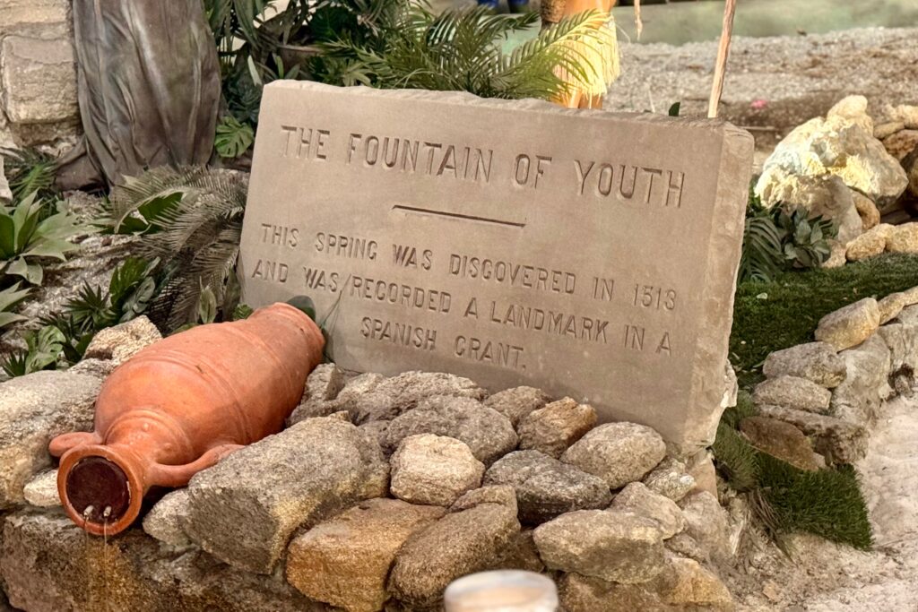 A large stone sign reads, "The Fountain of Youth". There is a clay cistern with water running out of it.  It is surrounded by rocks. 