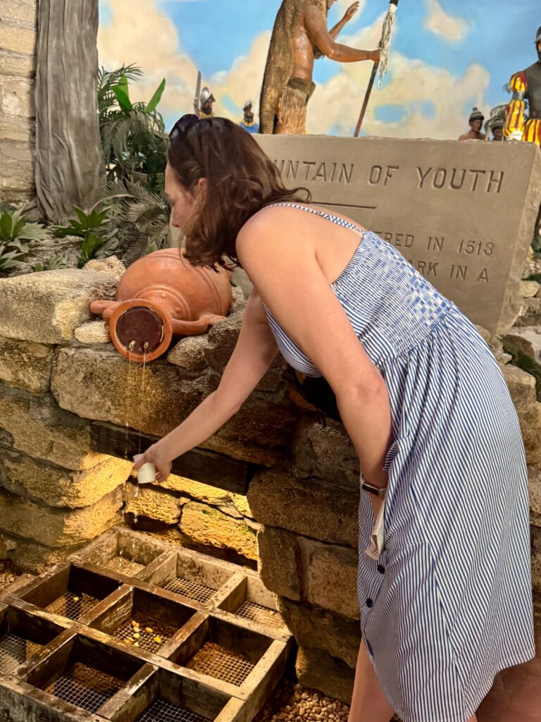 A woman in a dress bends slightly to fill a cup of water from a clay cistern.