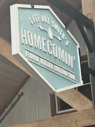 A wooden sign, painted blue and white, hangs from an L shaped bracket.  It reads, "Chef Art Smith's Homecomin', Florida Kitchen, Southern Shine."