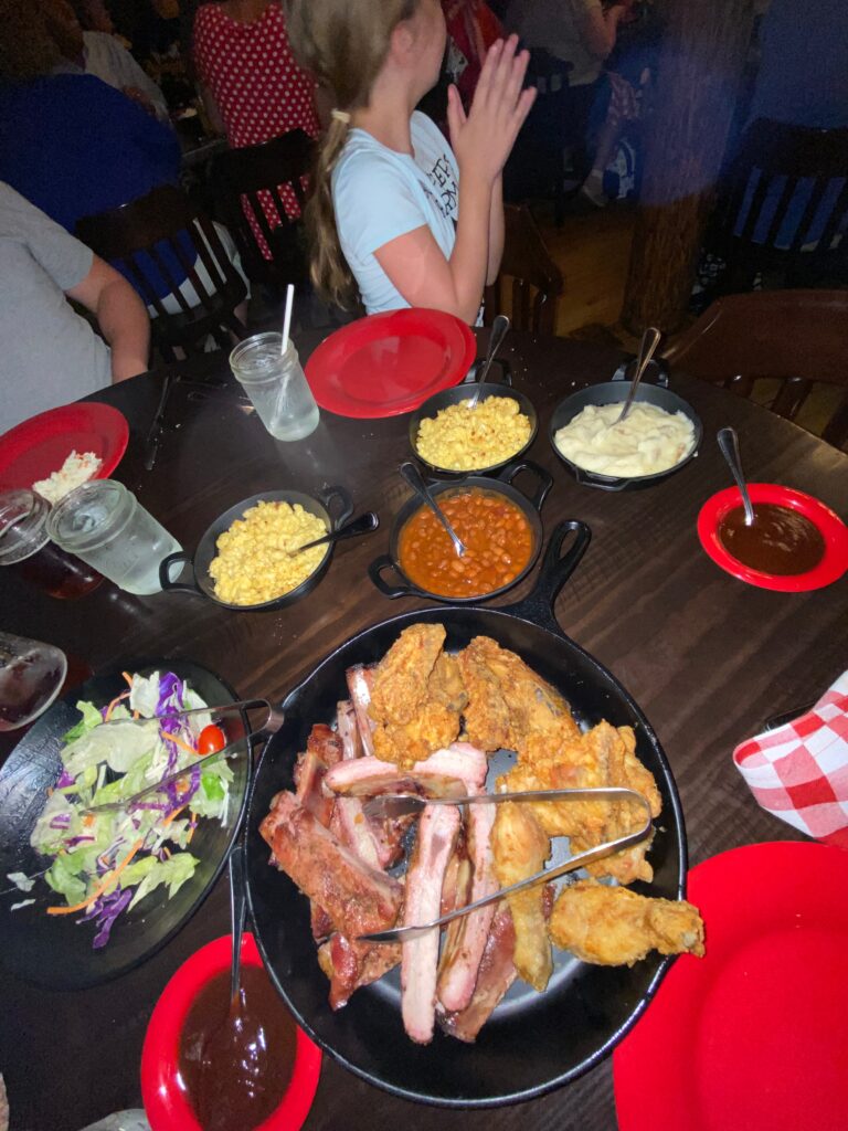 A table with several plates of food including: salad, macaroni and cheese, baked beans, fried chicken, mashed potatoes and ribs on the bone.  There are red checked cloth napkins on the table as well as a few water glasses. 