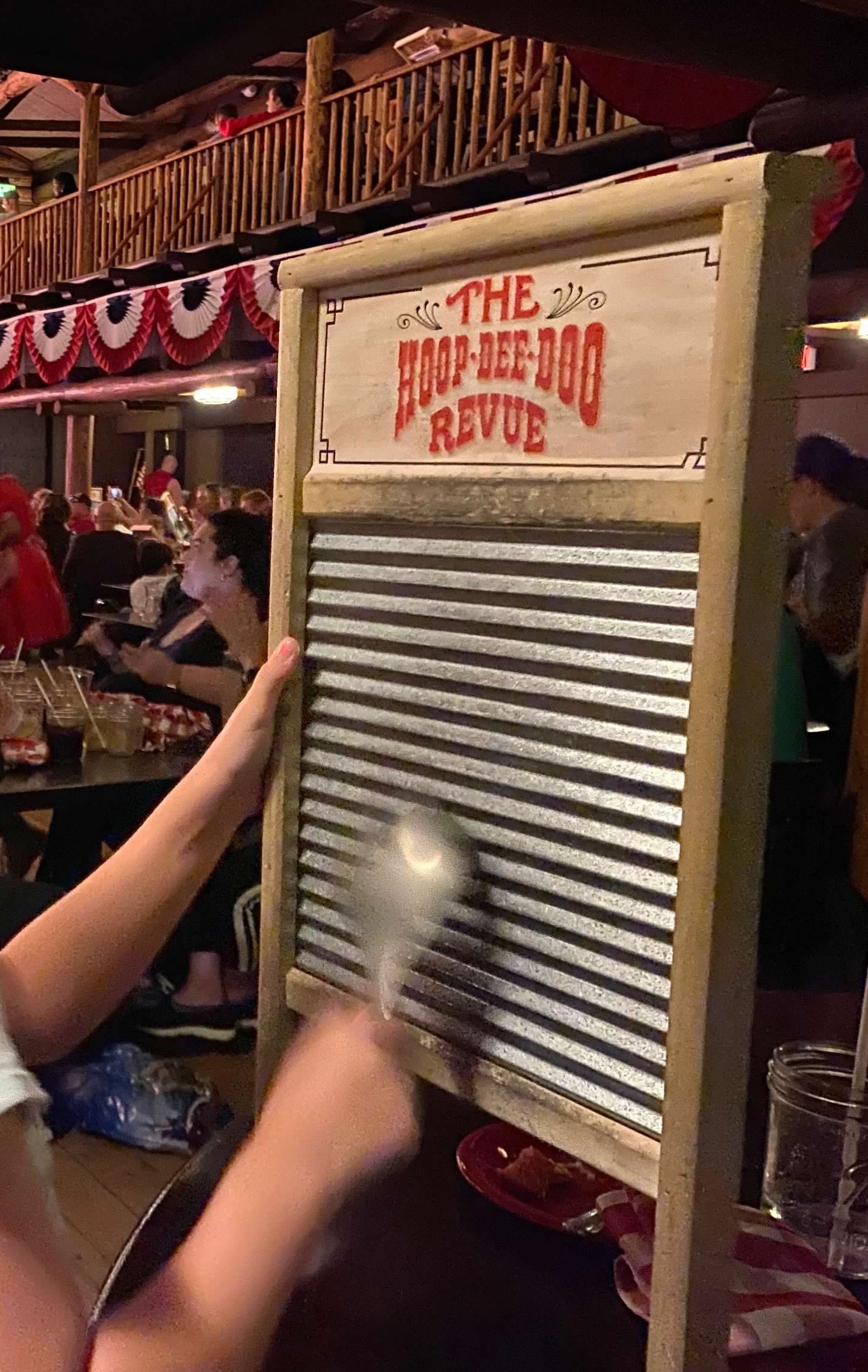 An old-fashioned washboard with the words, "Hoop-Dee-Doo Revue" in red font at the top.  A person is holding it and banging a spoon on it. 